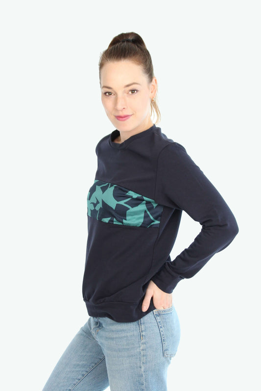 Eine seitlich stehende Frau, in einem dunkelblauen Pullover mit grün-blauen Muster auf der Brustpartie.