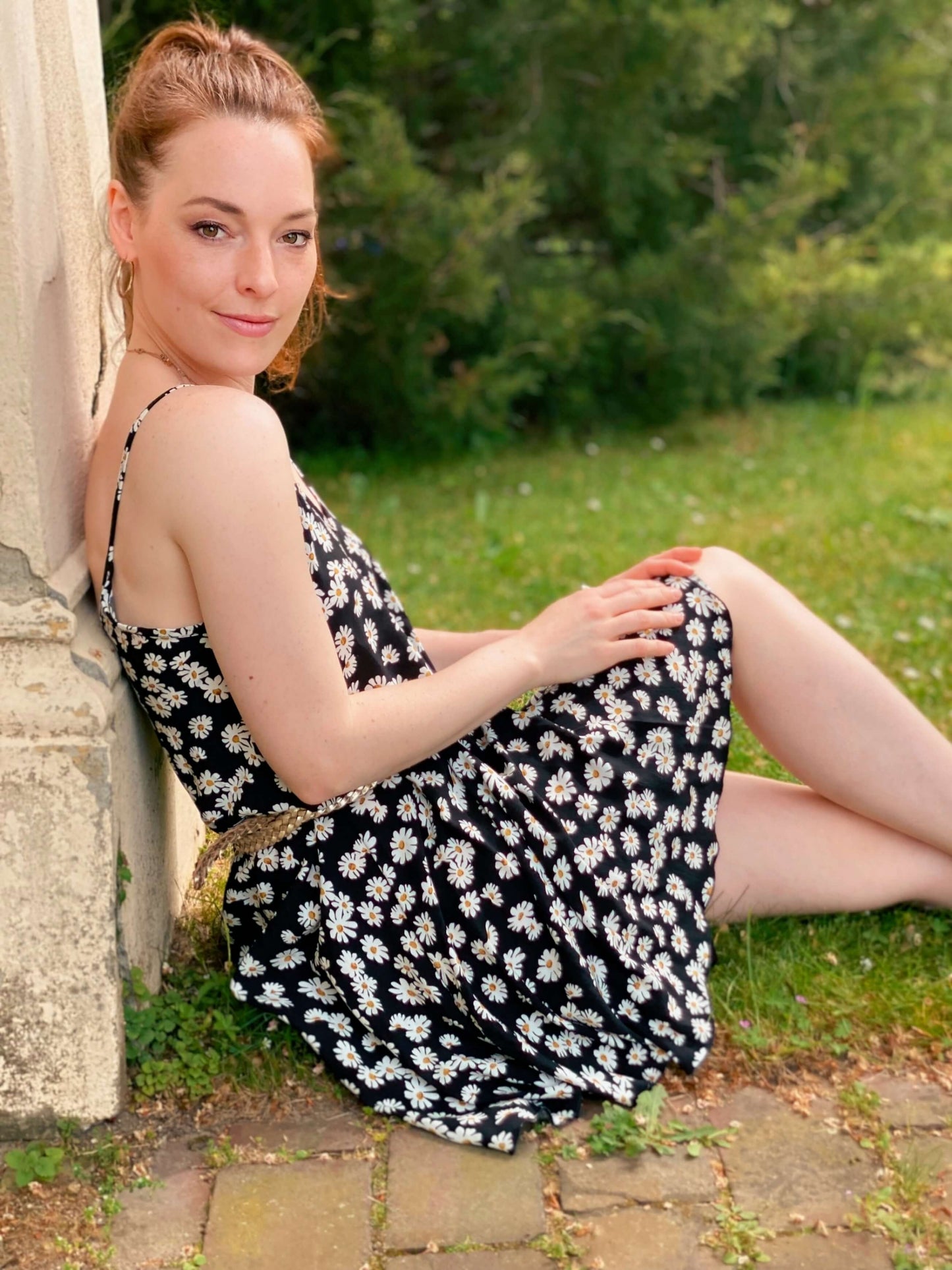 Frau, sitzt an einer Mauer gelehnt auf dem Boden. Sie trägt ein Sommerkleid mit Gänseblümchen Muster in schwarz und weiß.